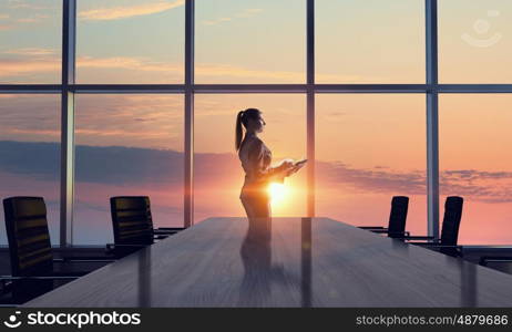 She is an early riser. Mixed media. Businesswoman in night office against panoramic window in light of sunset