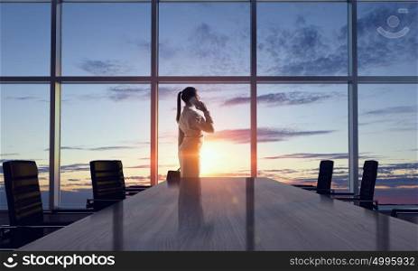 She is an early riser. Mixed media. Businesswoman in night office against panoramic window in light of sunset