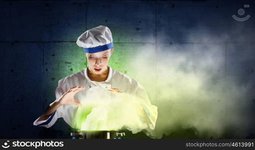 She is magician as cook. Pretty woman cook in hat and apron with doing magic above pot