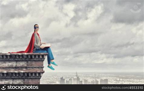She is super woman. Young thoughtful woman in red cape and mask on building roof read book