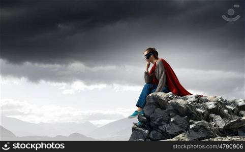 She is super woman. Young thoughtful woman in red cape and mask on rock top