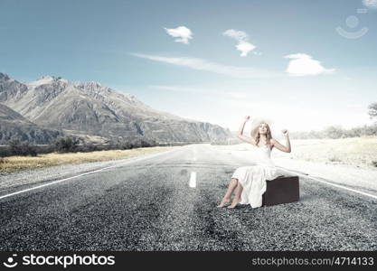 She is traveling light. Woman in white long dress and hat sitting on her luggage on asphalt road