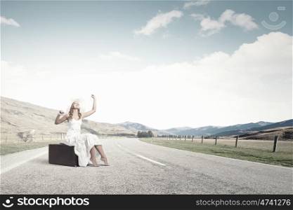 She is traveling light. Woman in white long dress and hat sitting on her luggage on asphalt road