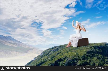 She is traveling light. Woman in white long dress and hat sitting on her luggage on mountain top