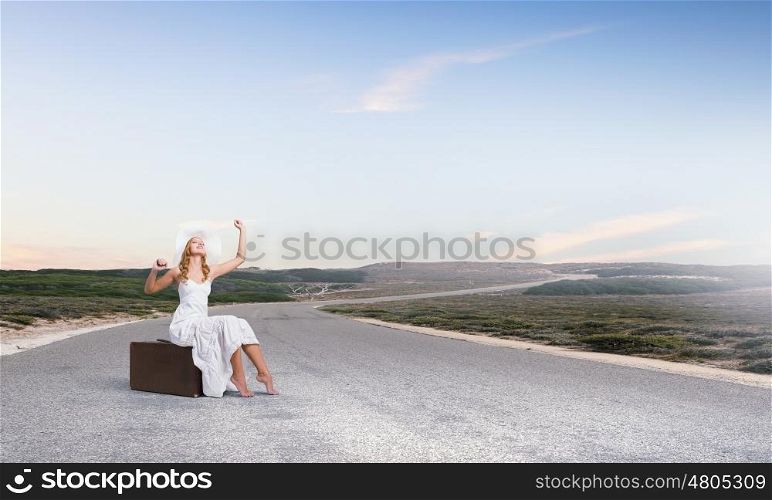 She is traveling light. Woman in white long dress and hat sitting on her luggage on asphalt road