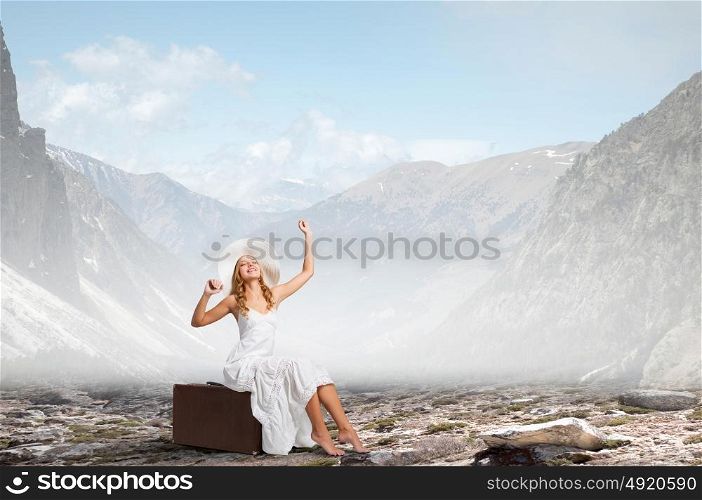 She is traveling light. Woman in white long dress and hat sitting on her luggage