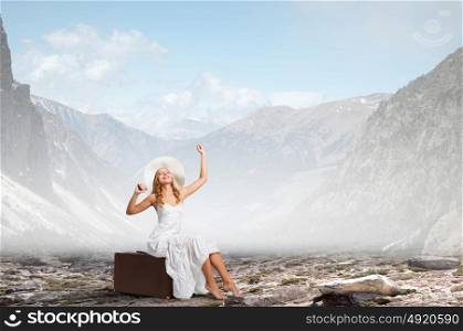 She is traveling light. Woman in white long dress and hat sitting on her luggage