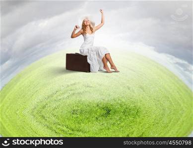 She is traveling light. Woman sitting on suitcase in white long dress and hat