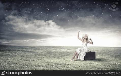 She is traveling light. Woman sitting on suitcase in white long dress and hat