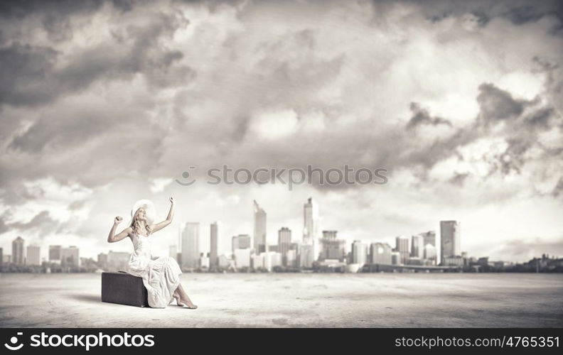 She is traveling light. Woman sitting on suitcase in white long dress and hat