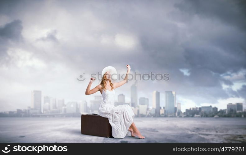 She is traveling light. Woman sitting on suitcase in white long dress and hat