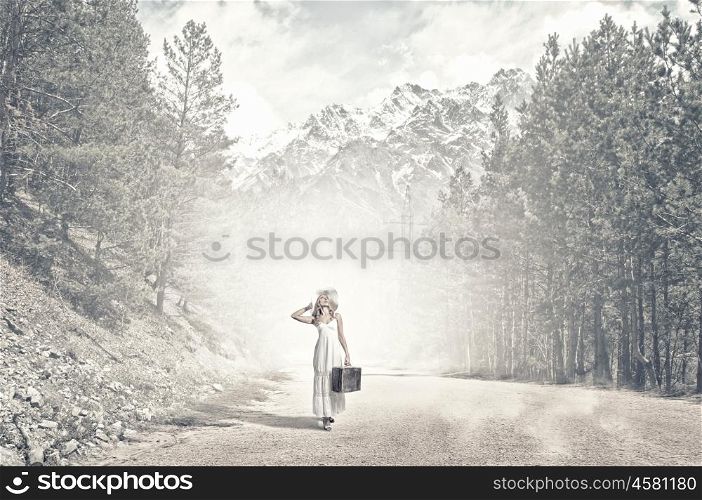 She is traveling light. Woman with suitcase in white long dress and hat on forest road