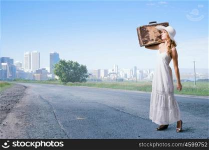 She is traveling light. Woman with suitcase in white long dress and hat on asphalt road