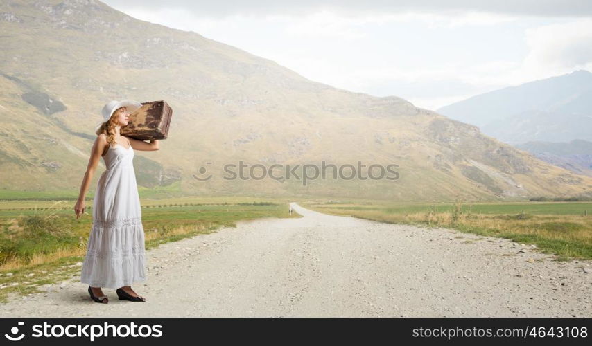 She is traveling light. Woman with suitcase in white long dress and hat on countryside road