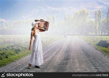 She is traveling light. Woman with suitcase in white long dress and hat on countryside road