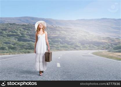She is traveling light. Woman with suitcase in white long dress and hat on asphalt road
