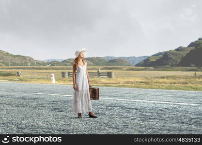 She is traveling light. Woman with suitcase in white long dress and hat on asphalt road