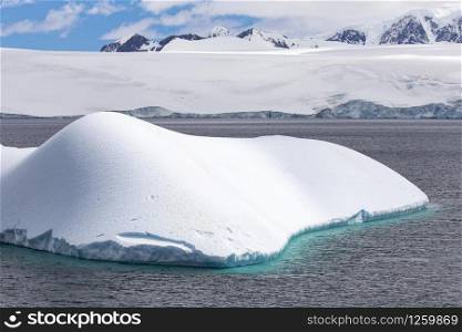Shimmering turquoise iceberg in front of mountains with glaciers in Antarctica