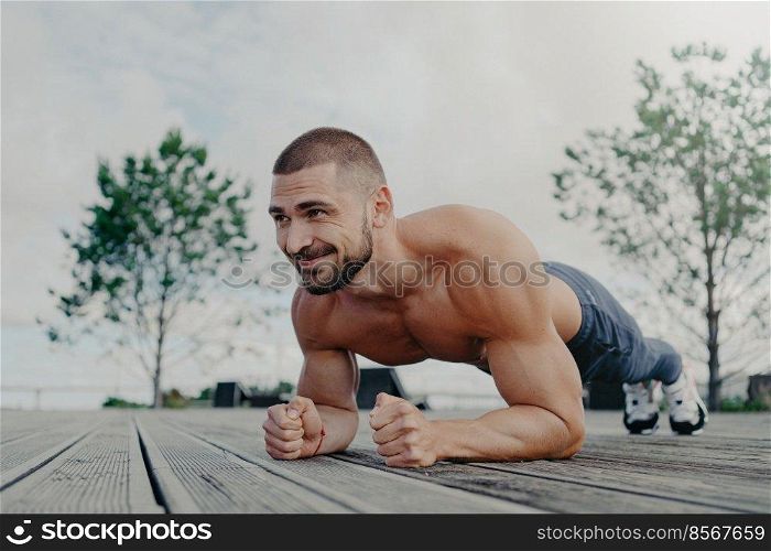 Shirtless determined bearded man with naked torso stands in plank pose and smiles gladfully, demonstrates endurance, poses outdoor, thinks about his body and health. Athletic guy trains outside
