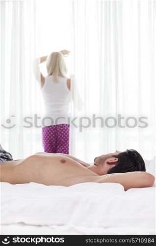 Shirtless young man sleeping in bed with woman standing by window