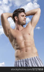 Shirtless young man with hands behind head against cloudy sky