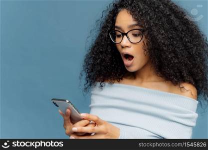 Shocked African American woman gots message from ex, gasps from surprise and excitement, reads stunning news on mobile phone, has curly bushy hair, wears jumper, isolated over blue studio wall