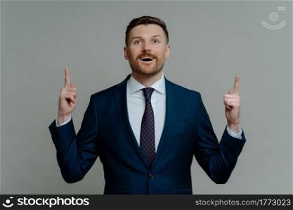 Shocked amazed business leader in suit pointing up with his fingers and feeling excited with open mouth while standing against grey background, male entrepreneur having great innovative idea. Shocked businessman in suit pointing up with fingers and looking with amazed face expression