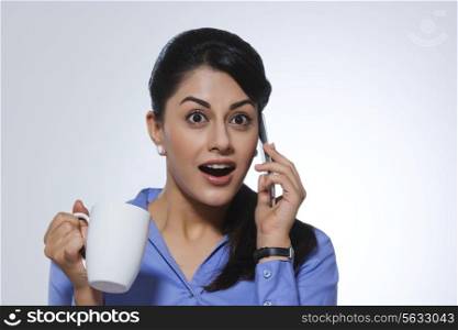 Shocked businesswoman with coffee mug answering call against gray background