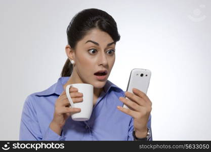 Shocked businesswoman with coffee mug reading text message on smart phone against gray background