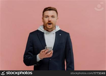 Shocked red haired guy reading unbeliveble news on smartphone isolated over pastel pink background. Young emotional man in casual clothes looking at camera with surprised face expression. Young excited man holding smartphone and looking at camera with surprised face expression