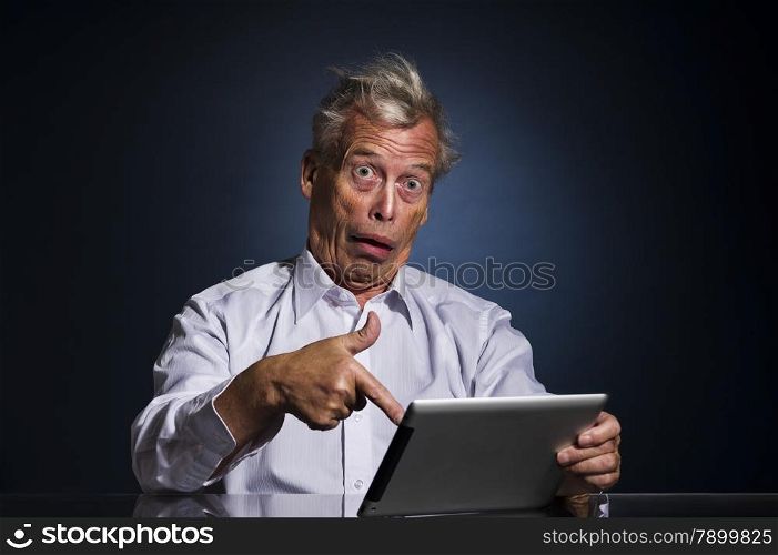 Shocked senior man pointing to his tablet computer with his finger with a look of appalled disbelief and confusion, humorous upper body studio portrait