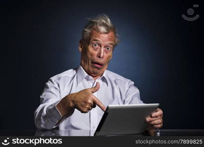 Shocked senior man pointing to his tablet computer with his finger with a look of appalled disbelief and confusion, humorous upper body studio portrait