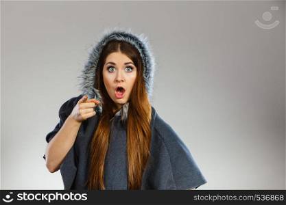 Shocked woman poitning at camera wearing dark poncho with furry hood. Winter fashion, trendy clothing outfits concept.. Shocked woman wearing dark poncho with hood