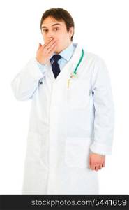 Shocked young medical doctor holding hand near mouth isolated on white&#xA;