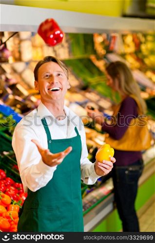 Shop assistant in a supermarket at the vegetable shelf; he is tossing a bell pepper