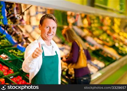 Shop assistant in a supermarket at the vegetable shelf; in the background a woman choosing vegetables