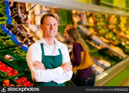 Shop assistant in a supermarket at the vegetable shelf; in the background a woman choosing vegetables