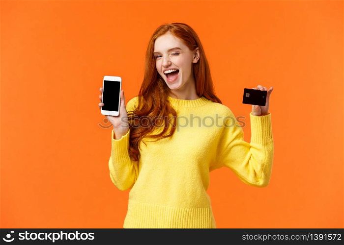 Shopping, advertising and mobile addication concept. Attractive cheeky redhead girl showing smartphone and credit card, wink and smile camera, promote online banking, deposit or payment method.. Shopping, advertising and mobile addication concept. Attractive cheeky redhead girl showing smartphone and credit card, wink and smile camera, promote online banking, deposit or payment method
