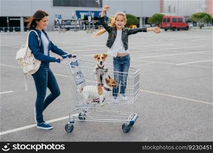 Shopping and family concept. Glad female carries trolley in which little girl stands with dogs, pose against shopping mall background, rejoice new purchases. People, happiness, day off concept
