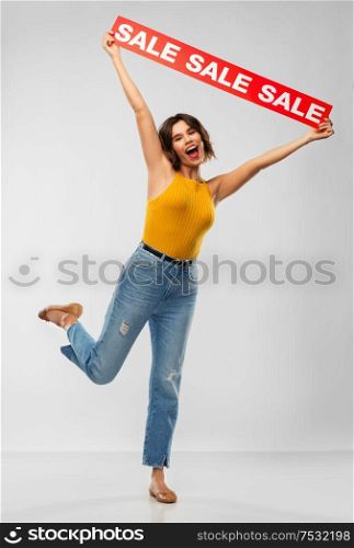 shopping and people concept - happy smiling young woman in mustard yellow top and jeans with sale banner posing over grey background. happy smiling young woman posing with sale banner