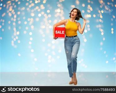 shopping and people concept - happy smiling young woman in mustard yellow top and jeans with sale sign posing over holidays lights on blue background. happy smiling young woman posing with sale sign
