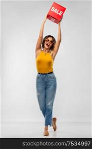 shopping and people concept - happy smiling young woman in sunglasses, mustard yellow top and jeans with sale sign posing over grey background. happy smiling young woman posing with sale sign