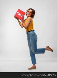 shopping and people concept - happy smiling young woman in sunglasses, mustard yellow top and jeans with sale sign posing over grey background. happy smiling young woman posing with sale sign