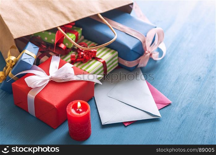 Shopping bag full of colorful presents, paper wrapped and tied with ribbon and bows, overturned on a blue wooden table, over closed envelopes, near a red candle.