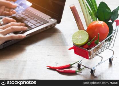 Shopping cart with full of fresh vegetables from market, Shopping online concept with woman using laptop and credit card in background