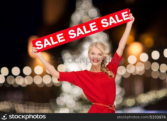 shopping, christmas and people concept - smiling woman in dress with red sale sign over lights background. woman in dress with red sale sign