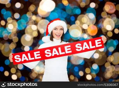 shopping, christmas and people concept - smiling woman in santa helper hat with red sale sign over lights background. happy woman in santa hat with christmas sale sign