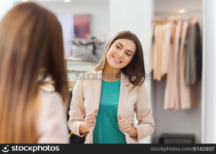 shopping, clothes, fashion, style and people concept - happy woman choosing jacket and posing at mirror in mall or clothing store