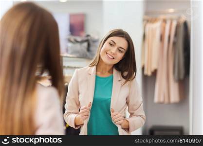 shopping, clothes, fashion, style and people concept - happy woman choosing jacket and posing at mirror in mall or clothing store