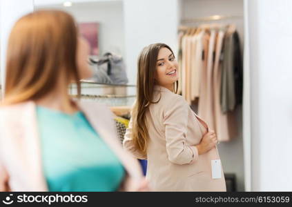 shopping, clothes, fashion, style and people concept - happy woman choosing jacket and posing at mirror in mall or clothing store